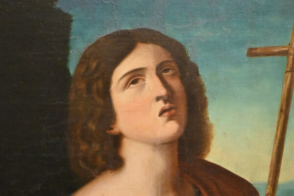 Pintura religiosa de San Juan Bautista con cruz de caña y túnica roja sobre fondo de paisaje, enmarcada en madera tallada.