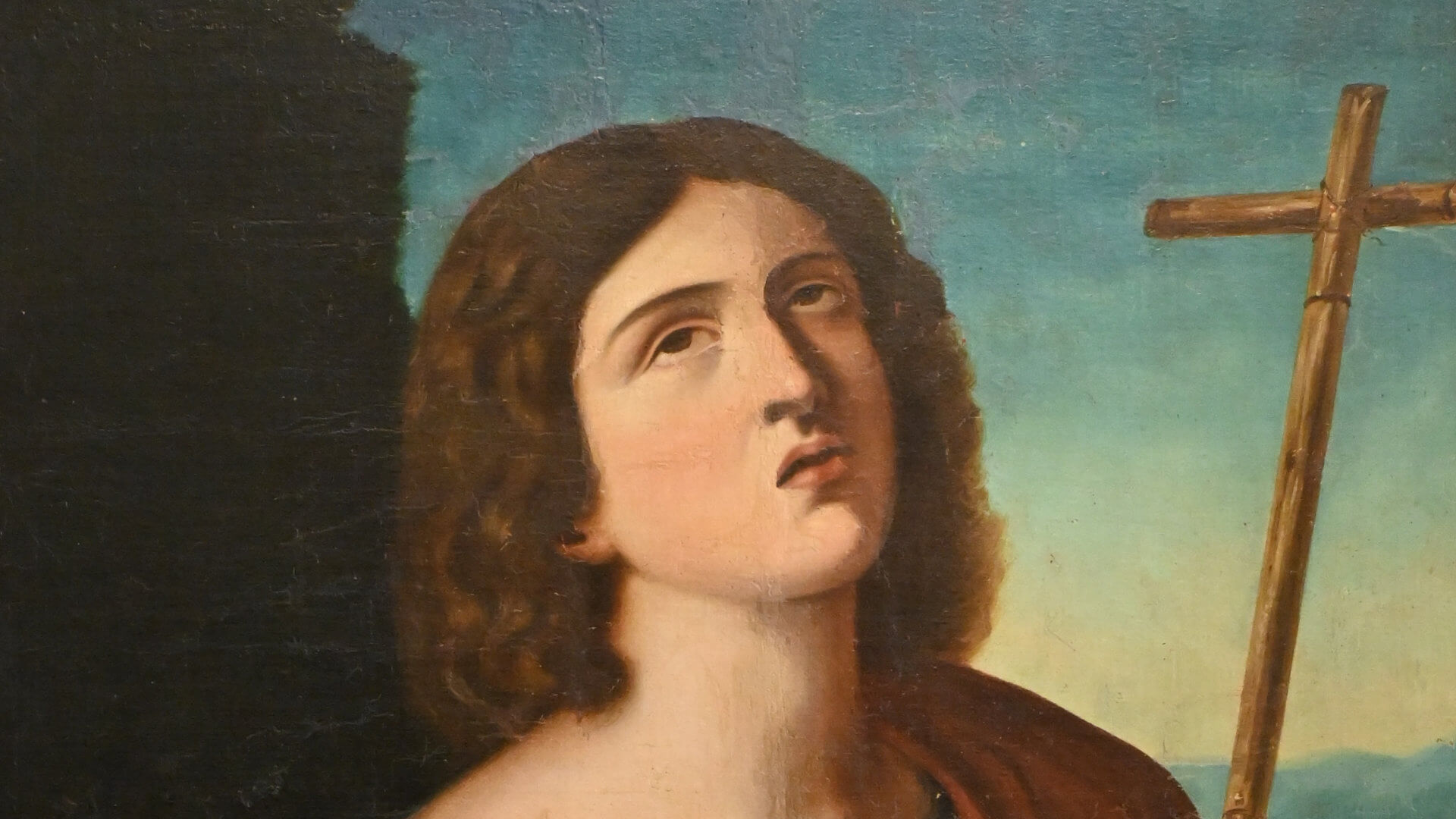 Pintura religiosa de San Juan Bautista con cruz de caña y túnica roja sobre fondo de paisaje, enmarcada en madera tallada.