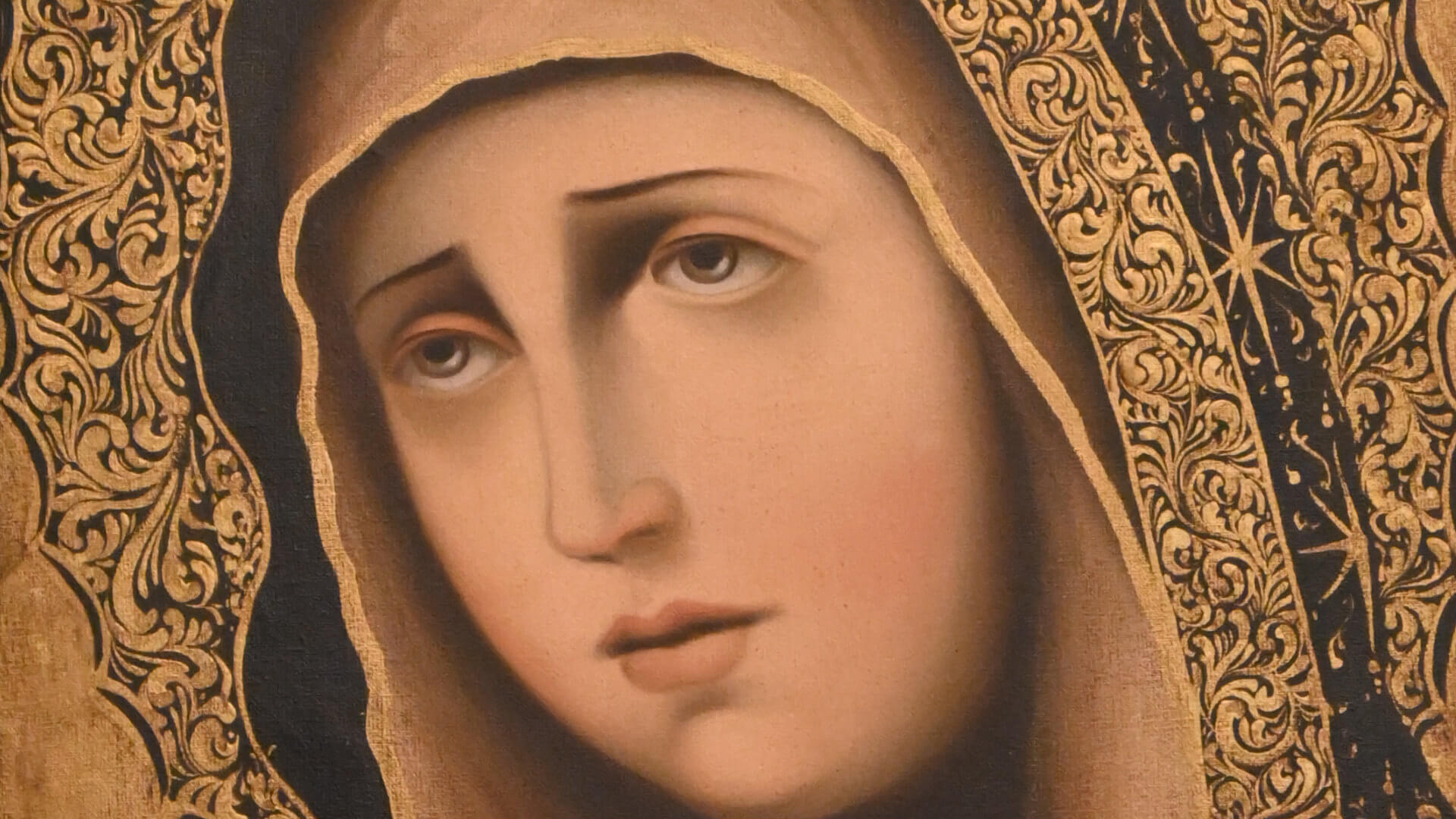 Pintura religiosa de la Virgen María con manto oscuro, halo dorado decorado y marco de madera tallada en tonos oscuros.